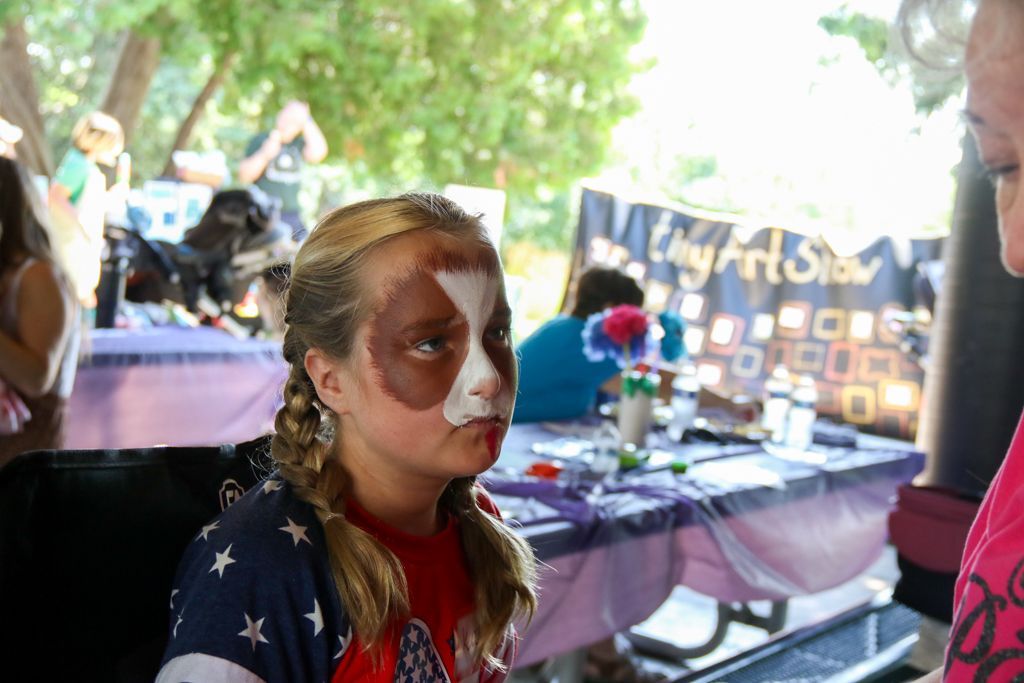 Girl with face paint, braids, and American flag shirt at a fair booth.
