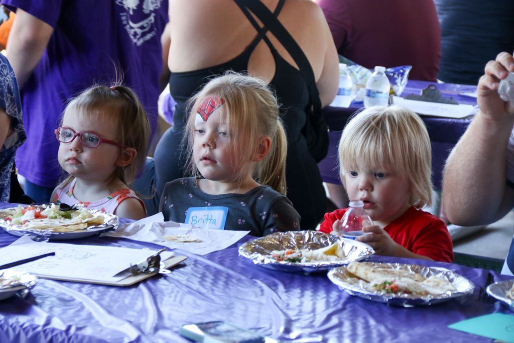 Three young children at a purple table eating. The middle child has a painted mark on their forehead.
