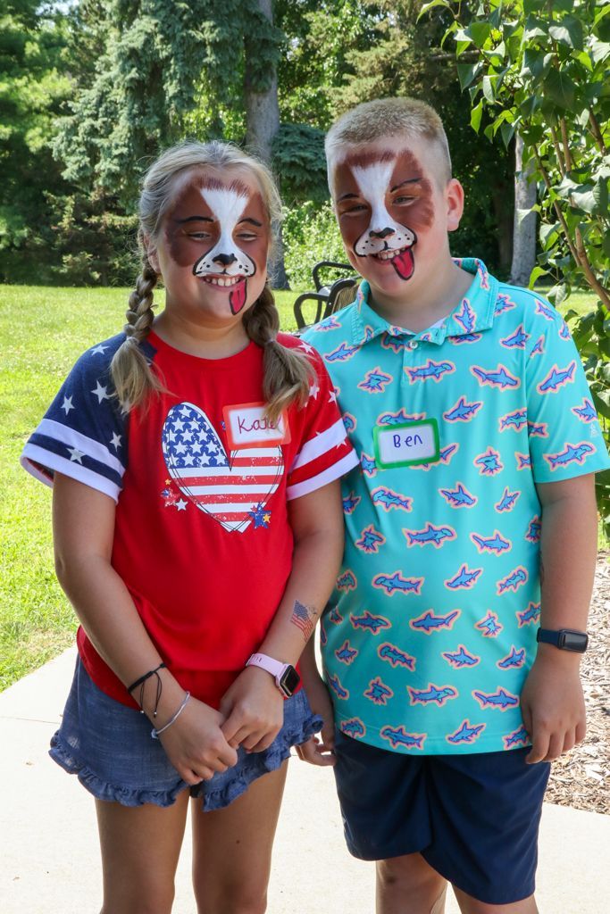 Two children with dog face paint smiling outdoors. The girl wears a red, white, and blue shirt. The boy wears a blue shirt.