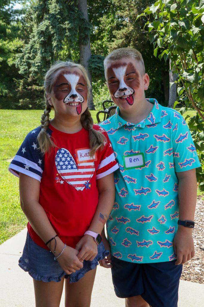 Two people with dog face paint smile outdoors. One wears a red and blue patriotic shirt, the other, a blue patterned shirt.
