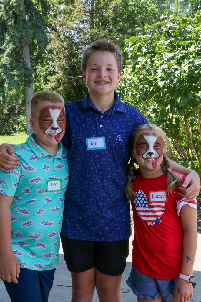 Three children smiling outdoors; two with dog face paint and one with arms around the others.
