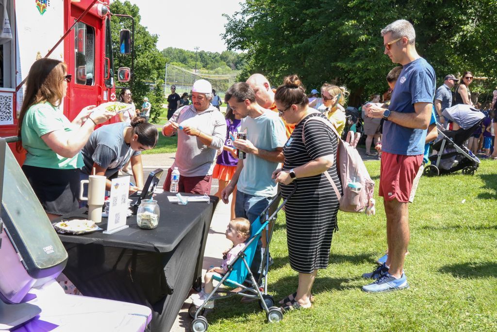 People lining up at a red food truck; a woman serves customers outdoors on a sunny day.