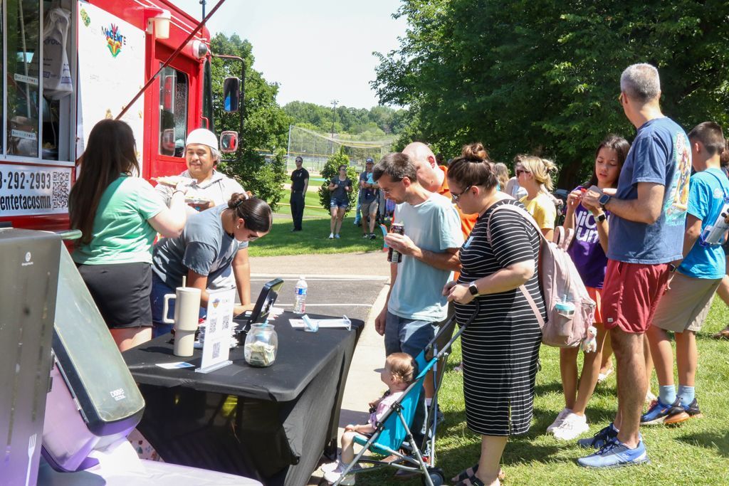 People line up to order food or drinks from a red food truck on a sunny day.