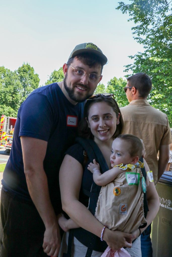 Family of three smiling, outdoors: Parents and baby in carrier. Man in hat, woman holding baby, another person in background.