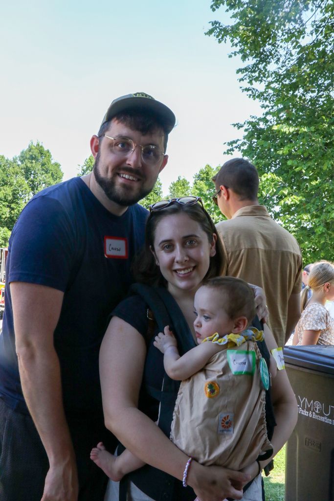 Family of three smiling outdoors: man with glasses, woman holding baby in carrier.