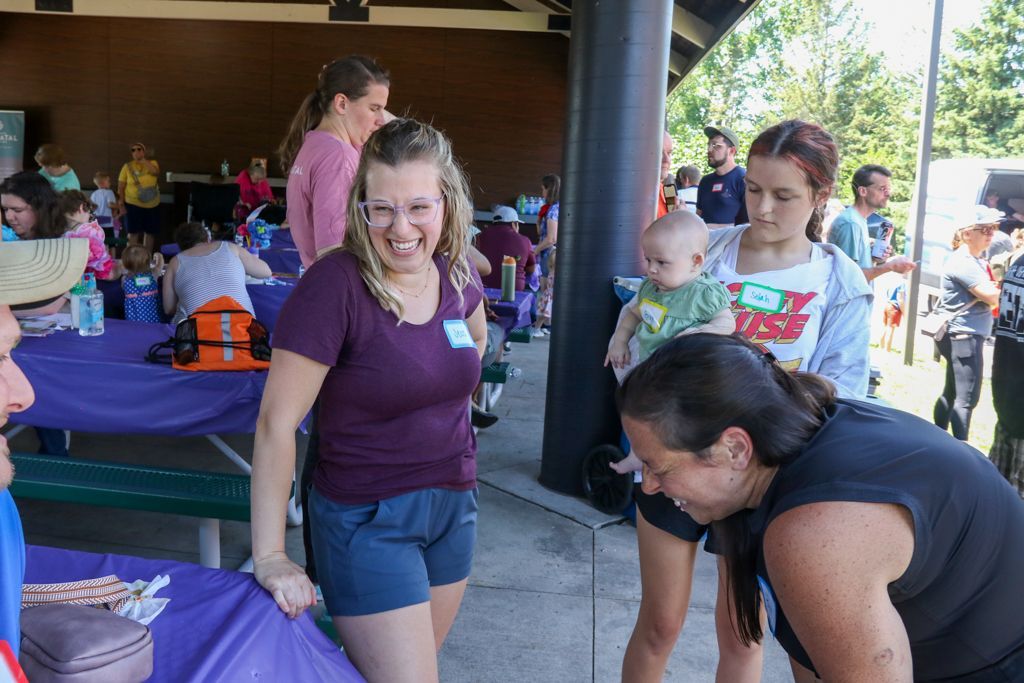 People at an outdoor event near a covered structure. Woman in purple shirt smiles. Others stand or sit at tables.
