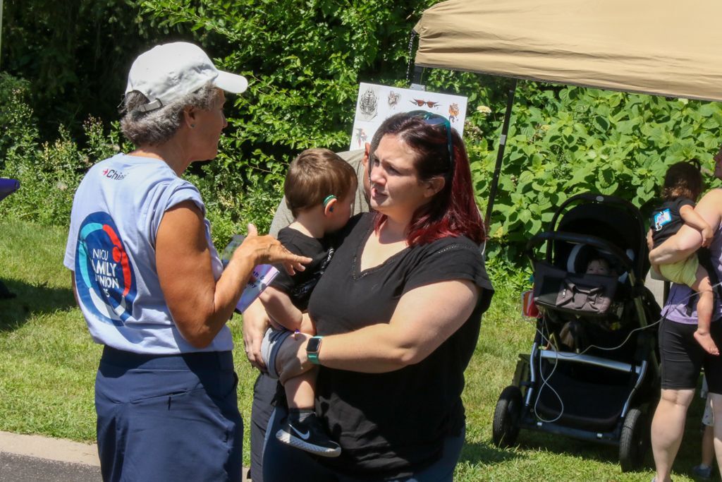 Woman in white hat speaks with another woman holding a child outdoors, a stroller is nearby.