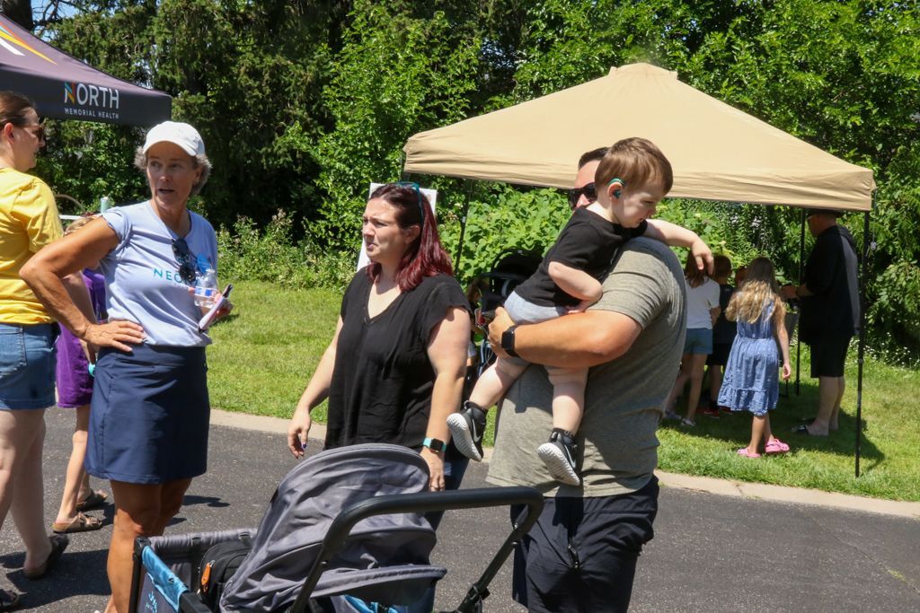 People at an outdoor event. Man holds child, woman pushes stroller. Tents and trees in the background.