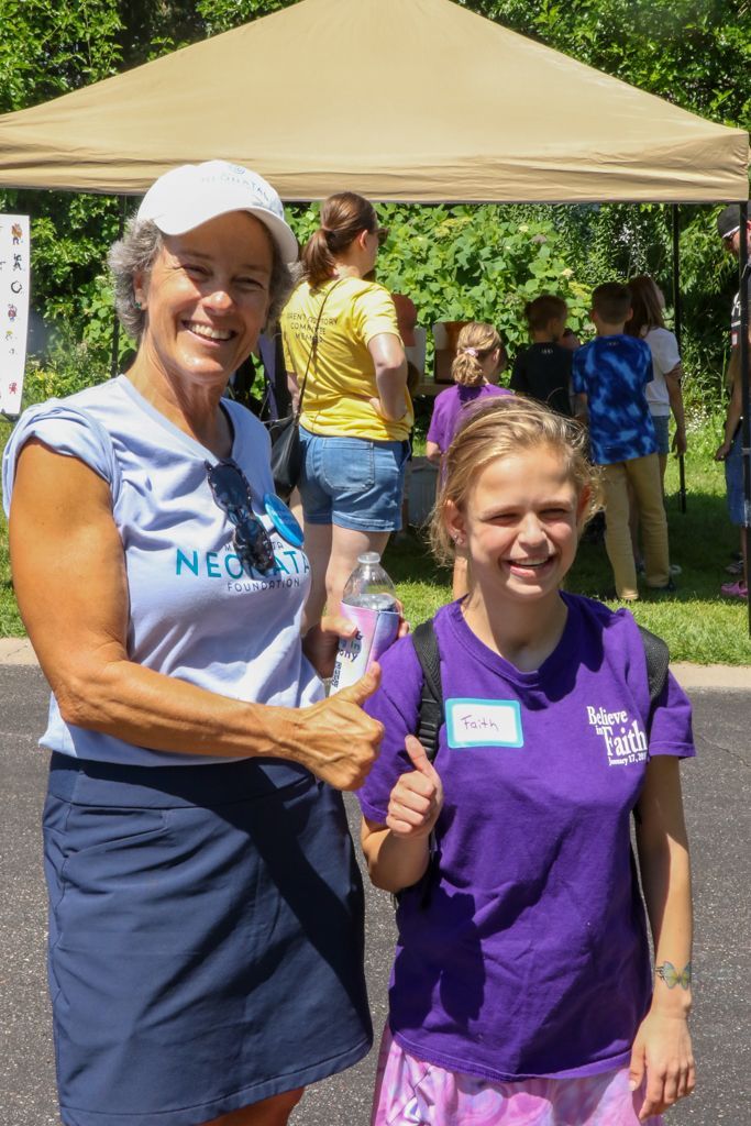 Woman in white hat and girl in purple shirt give thumbs up; outdoors, sunny day.