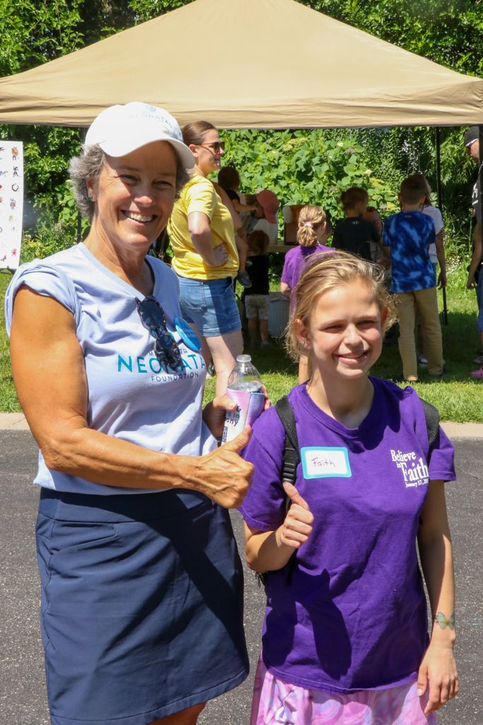 Two women smiling, giving thumbs-up. One in a white hat, light blue shirt, navy skirt; the other in a purple shirt, name tag. Outdoor setting.