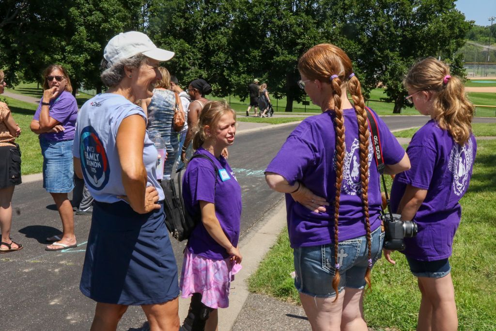 People in purple shirts and hats talking on a path outdoors in front of trees and a field.