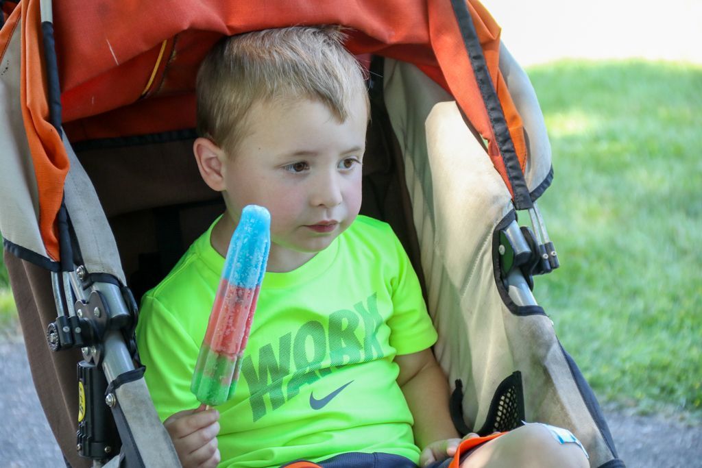 Boy in stroller holding a red, white, and blue popsicle, wearing a green shirt.