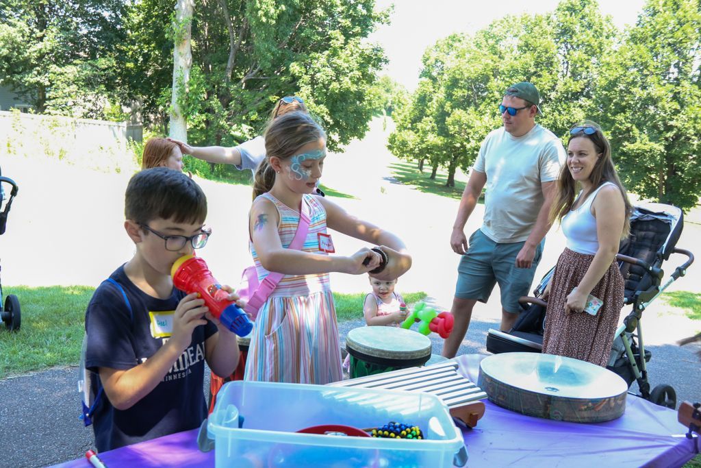 Children playing musical instruments at an outdoor event; adults watch nearby.