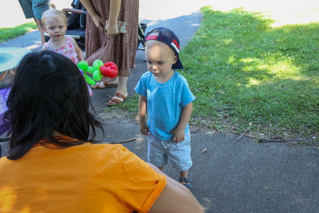 A young child in a blue shirt and baseball cap stands on a paved path, facing a person in an orange shirt.