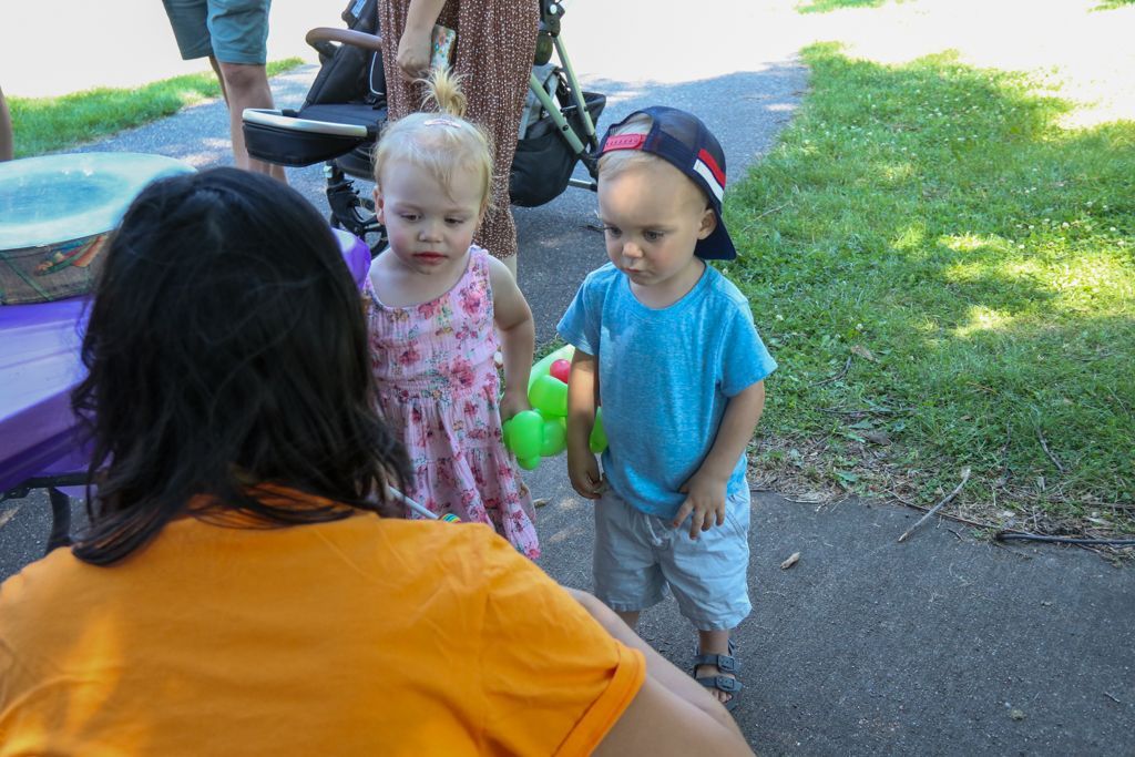 Two toddlers standing near a person in orange shirt outdoors; one holds a green toy.