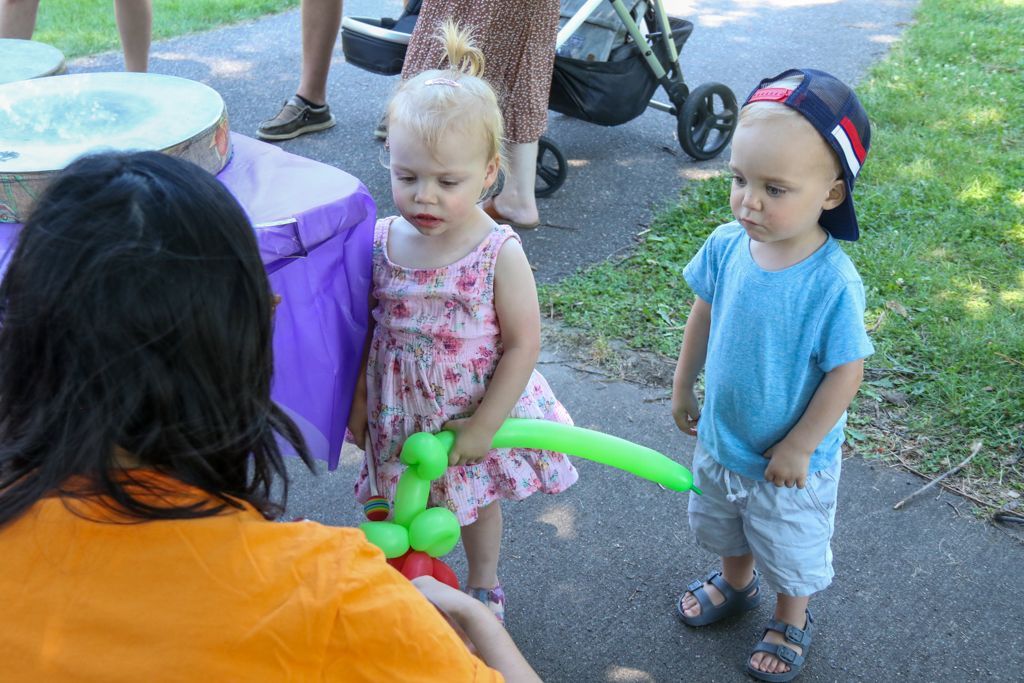 Two toddlers beside a person; one holds a balloon animal. Outdoor event with a table and stroller.