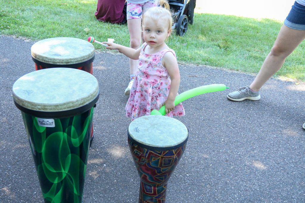 Young child in a pink dress holding a balloon and drumstick, playing drums outside.