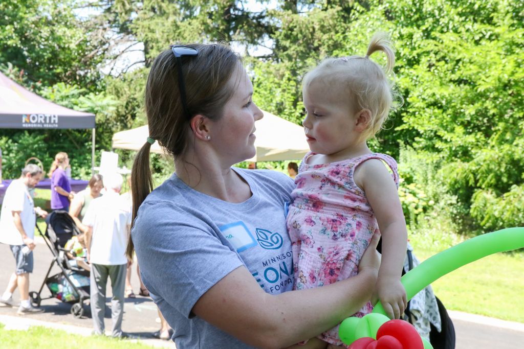 Woman holding a toddler outdoors; both smiling, greenery and event in background.