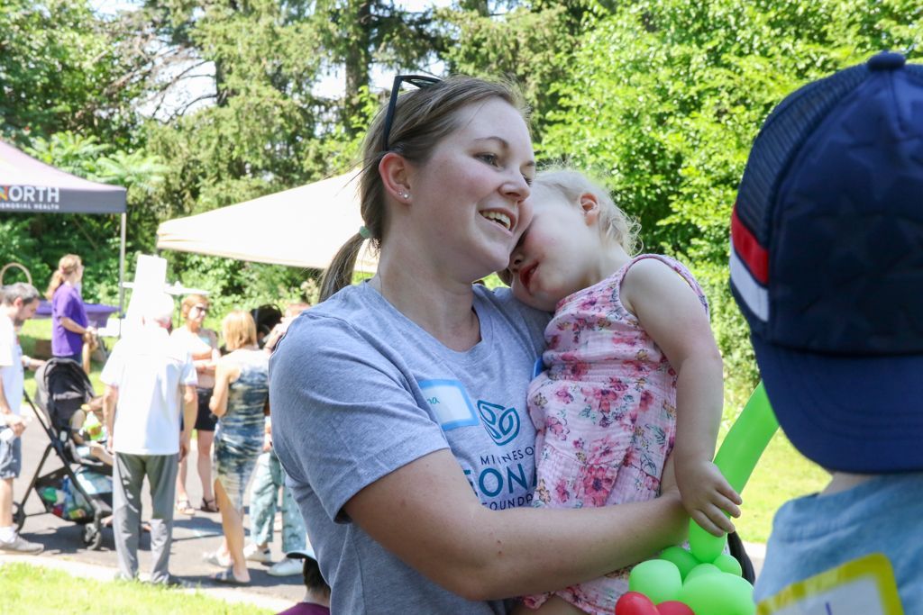 Woman smiles while holding a sleeping toddler outdoors; others in background.