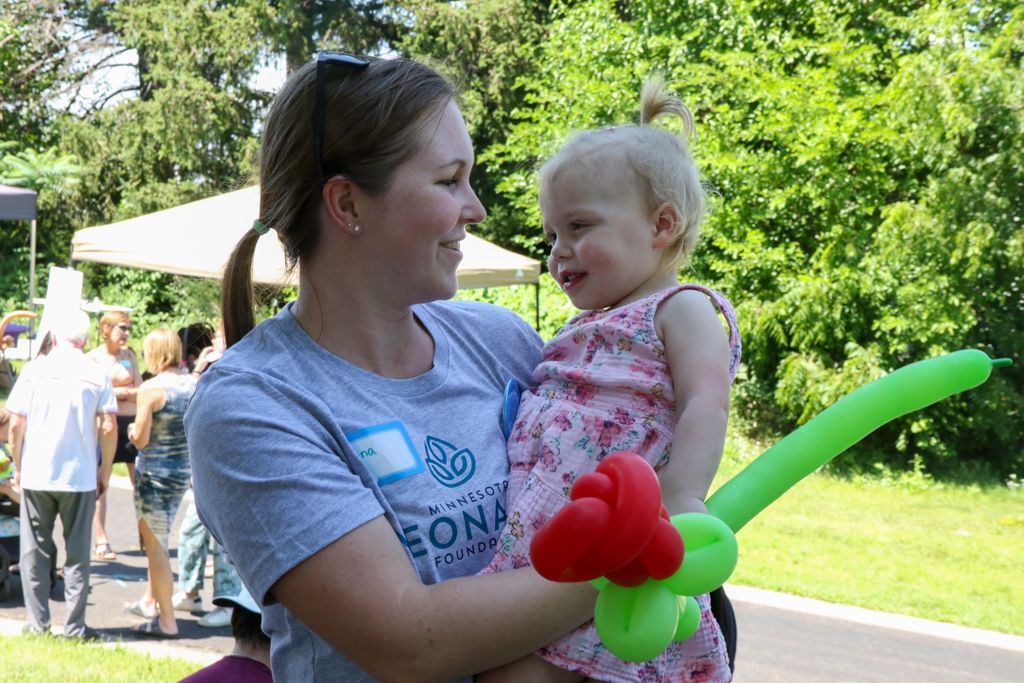 Woman holding a toddler at an outdoor event, looking at each other, green and red balloon nearby.