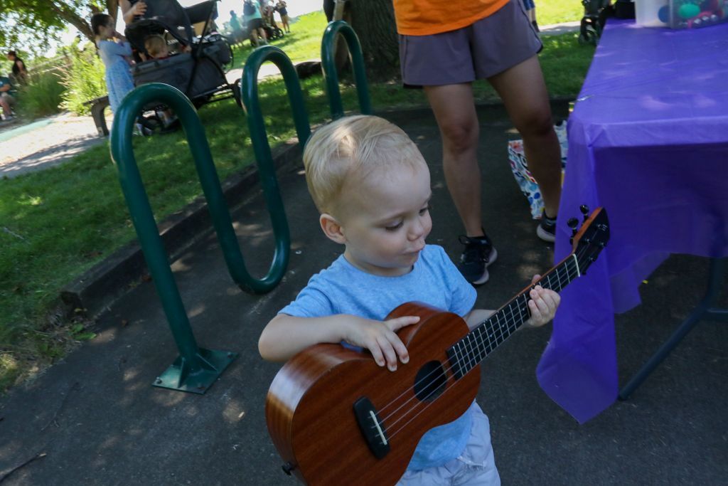 Young child playing a ukulele outdoors near a bike rack and purple table.