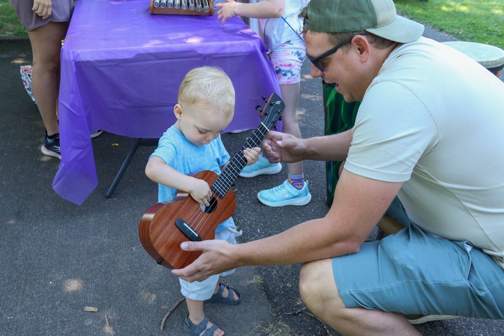 A toddler playing a ukulele, helped by an adult, outdoors near a purple table.