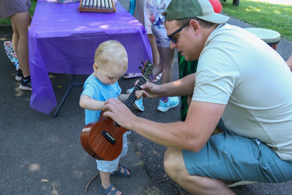 Man shows child how to hold a ukulele at an outdoor event with a purple tablecloth.
