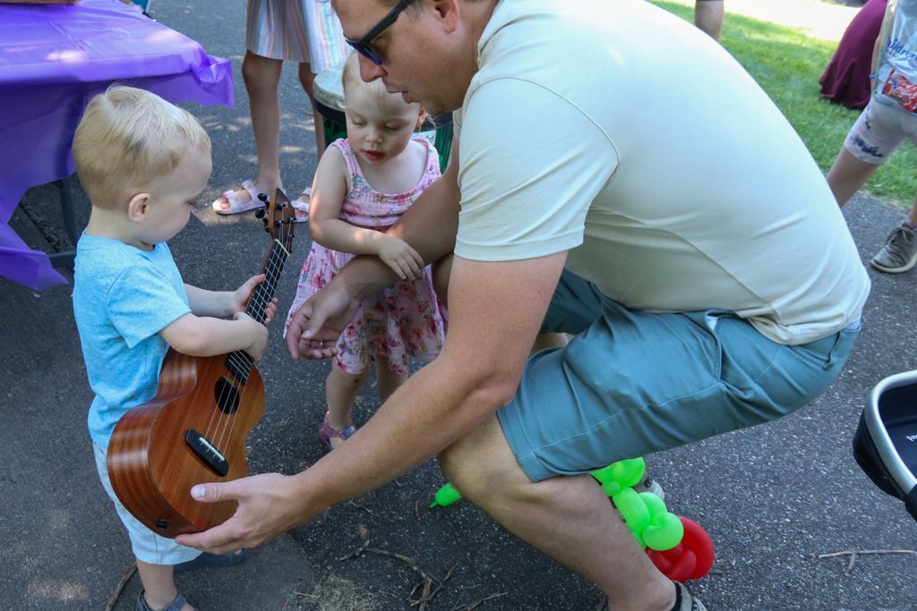 Man showing a toddler how to hold a ukulele outside; a child watches.