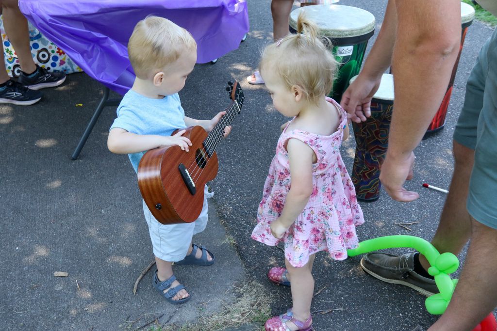 Two toddlers, boy with ukulele, girl in floral dress, looking at each other outdoors near drums, purple table.