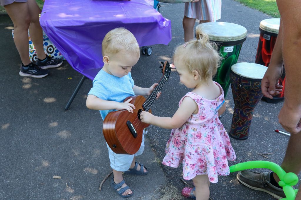 Two toddlers, one in blue, the other in pink, share a ukulele outdoors near drums.
