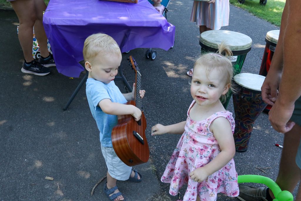 Two toddlers with a ukulele, purple table, and drums outdoors. One child holds the instrument, the other smiles.