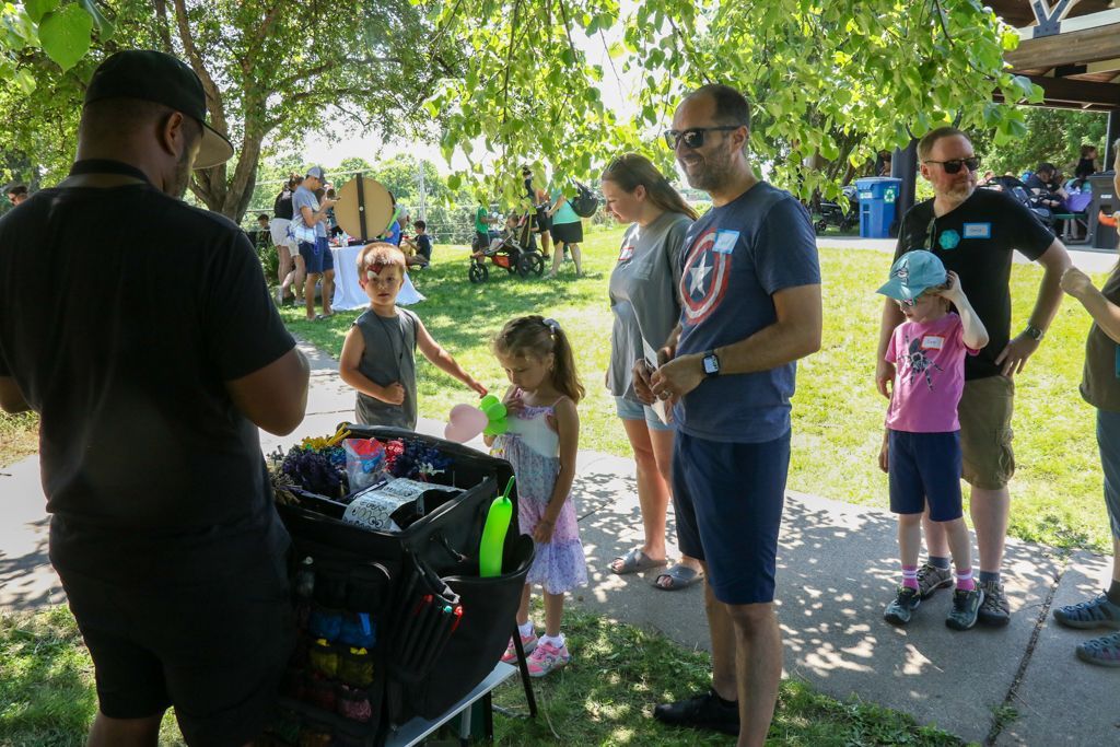 People gathered outdoors near a table with supplies; children and adults look on. Sunlight and trees are visible.