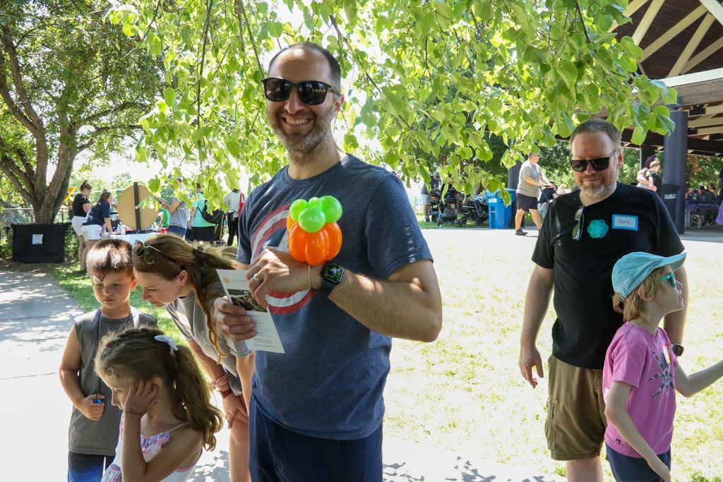 Man holding balloon smiles, children watch. Outdoor event, sunny day.