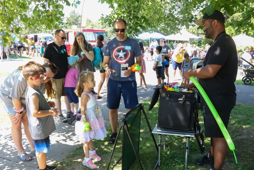 A balloon artist at an outdoor event makes a green balloon animal for children, who watch with anticipation.