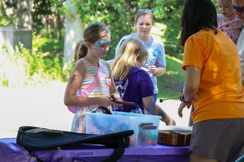 Children with face paint and a woman with an orange shirt at a table with musical instruments outside.