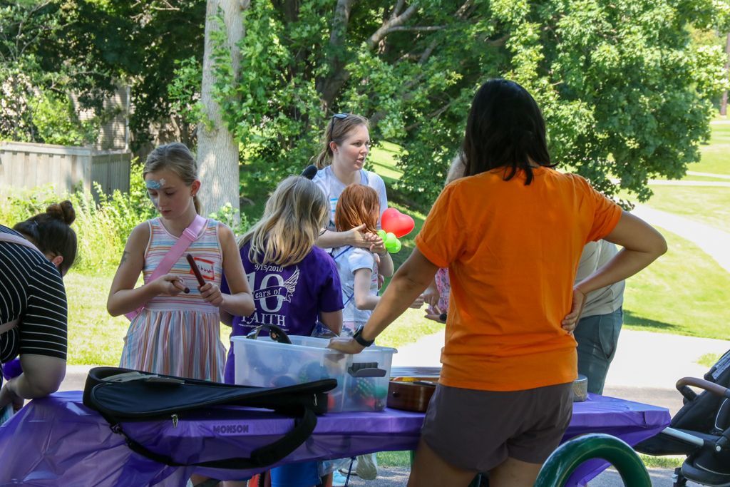 People at a table doing crafts outdoors; woman in orange shirt, kids with face paint.