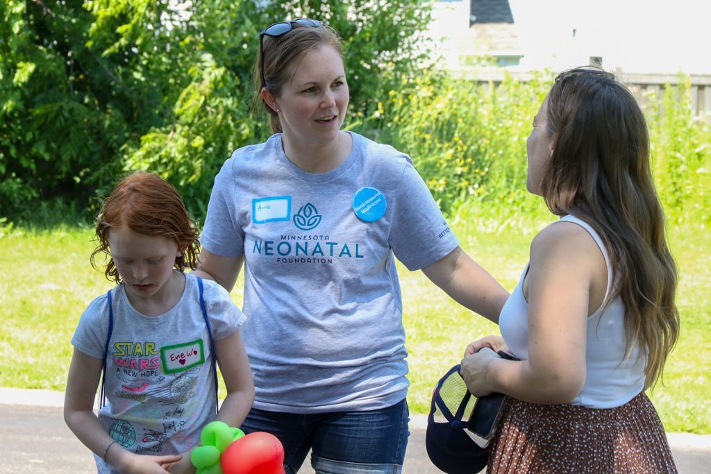 Three people outside. A woman in a grey t-shirt talks to another woman as a young girl stands nearby with balloons.