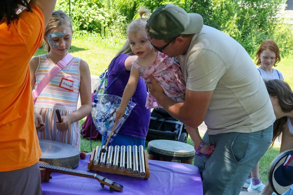 People at an outdoor event with musical instruments. A person holds a toddler; children play drums and look on.