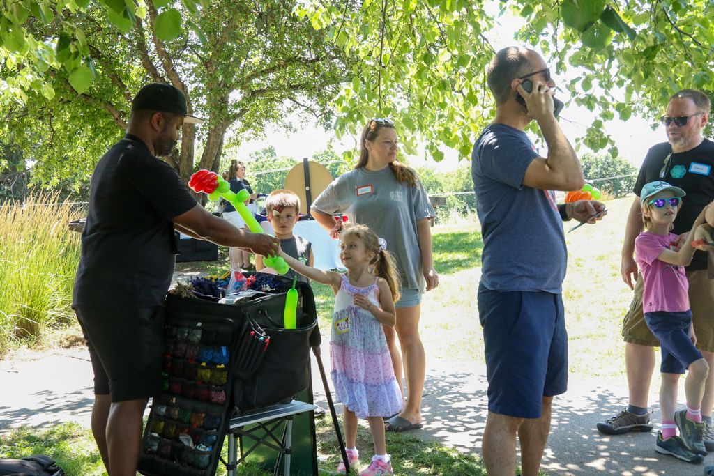People at an outdoor event, including a balloon vendor, a child interacting with the vendor, and adults.
