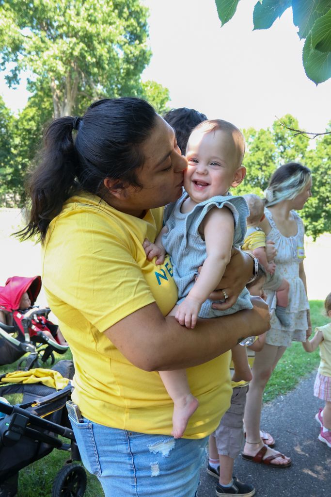 Woman in yellow shirt kisses a smiling baby outdoors. Green trees in the background.
