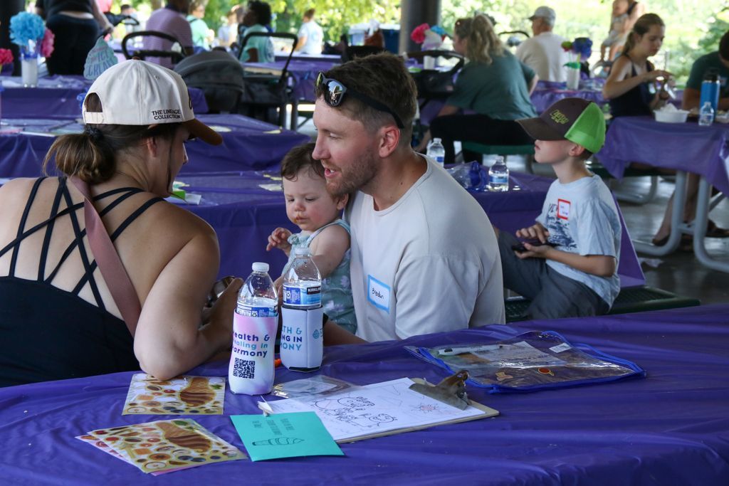 Family at picnic table: Father holding baby, child, and woman with hat; purple table cloth, sunny day.