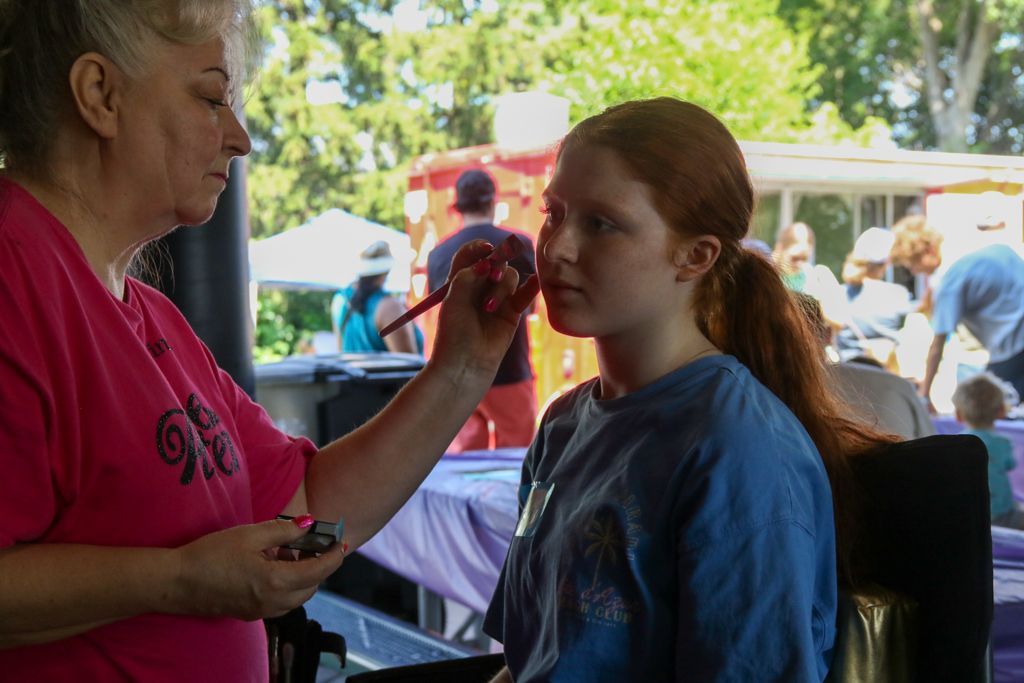 Woman applying face paint to a person with red hair. Outdoor event, people in background.