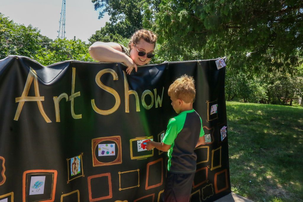 An art show banner with a young person placing artwork, an adult looking on. Outdoors, sunny.