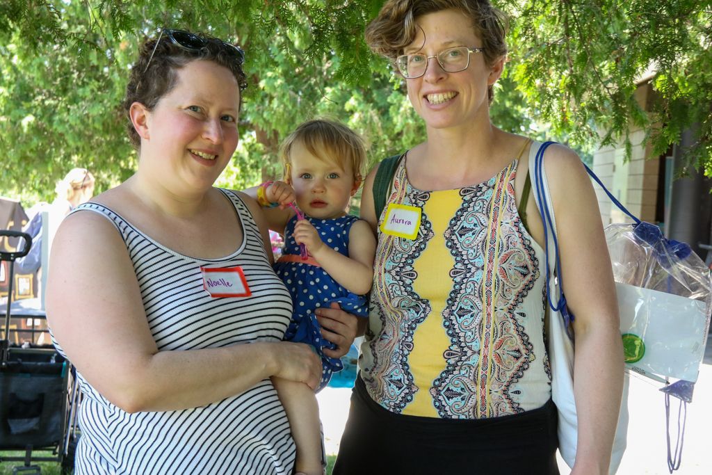 Two women and a child outdoors, smiling. One woman holds the child, the other wears a name tag.