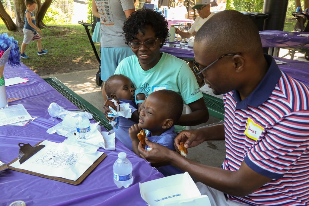 Family at outdoor event; parents feeding two babies at a purple table.