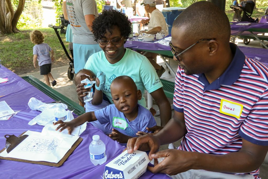 Family at a picnic table with children; an adult man opens a box while the others watch.