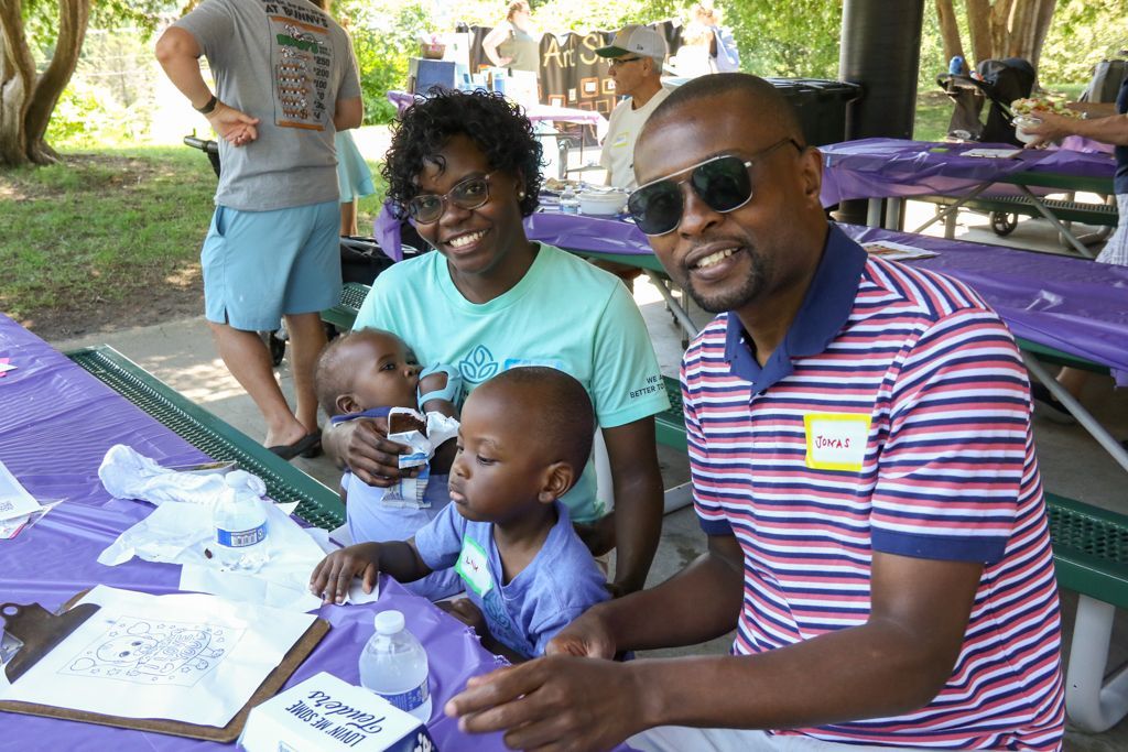 Family at a table, smiling. Woman holds a baby; a toddler sits nearby. Man in sunglasses, picnic setting.