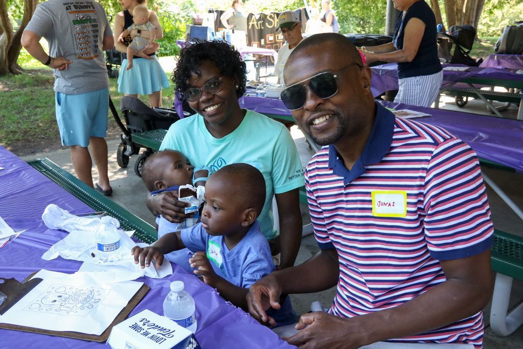 Family at picnic table smiling, with two babies, surrounded by other people, purple tablecloth.