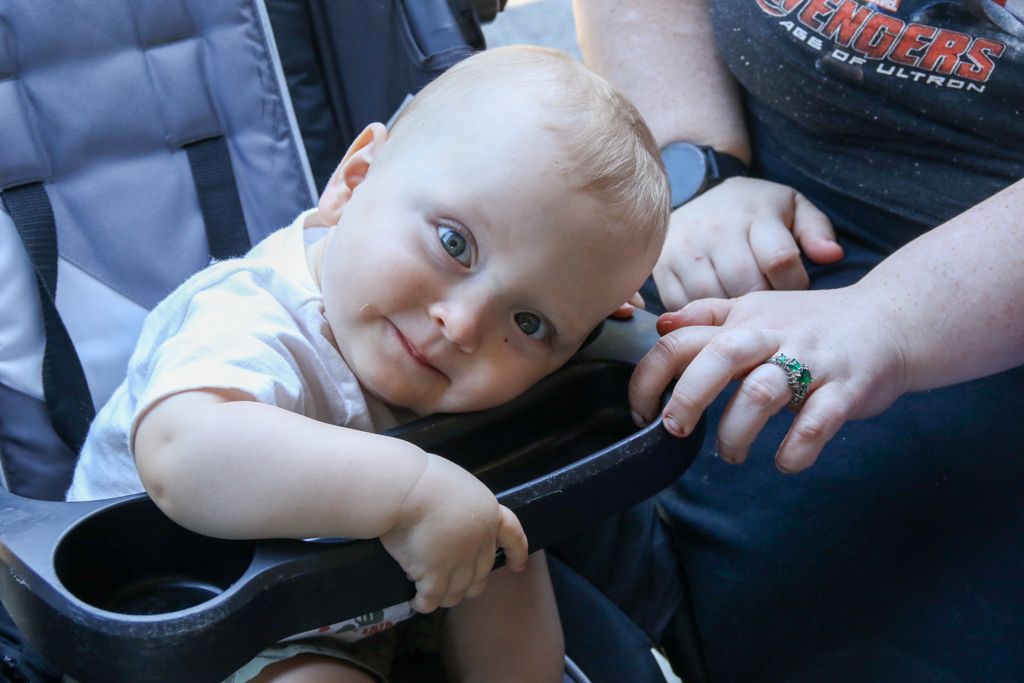 Baby in stroller looking at camera, leaning on the stroller bar with a slight smile, next to a person's arm.