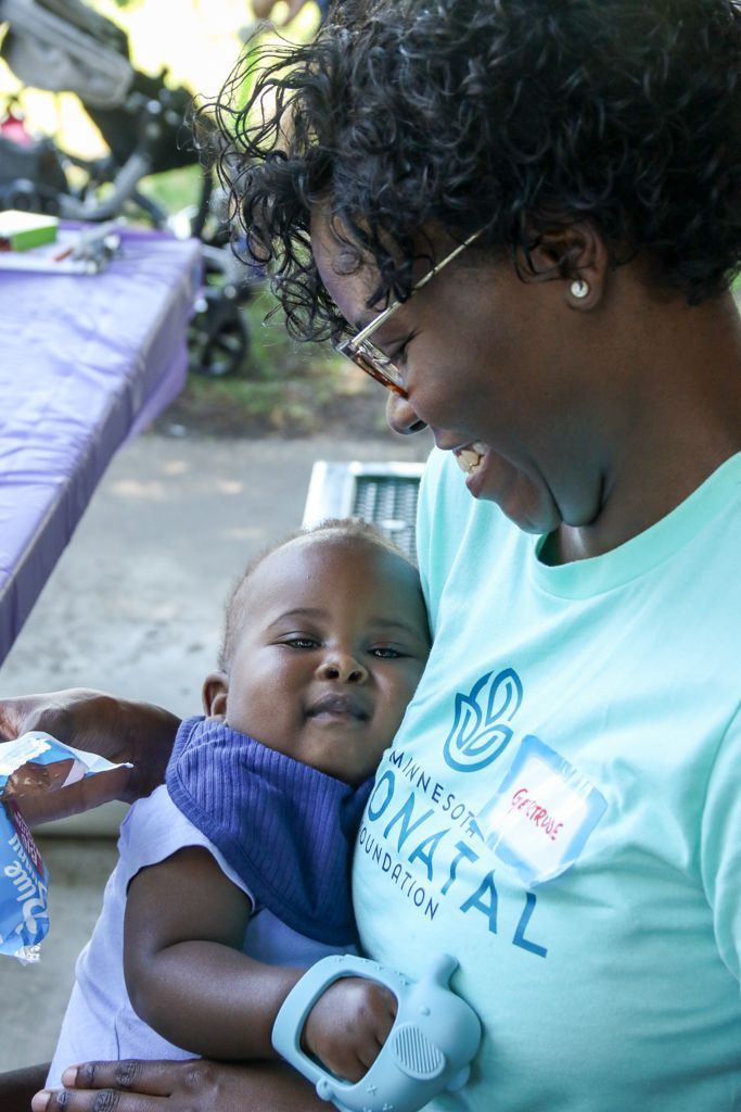 Woman holding baby, smiling. Both are outdoors. The woman wears a teal shirt with a logo and glasses.